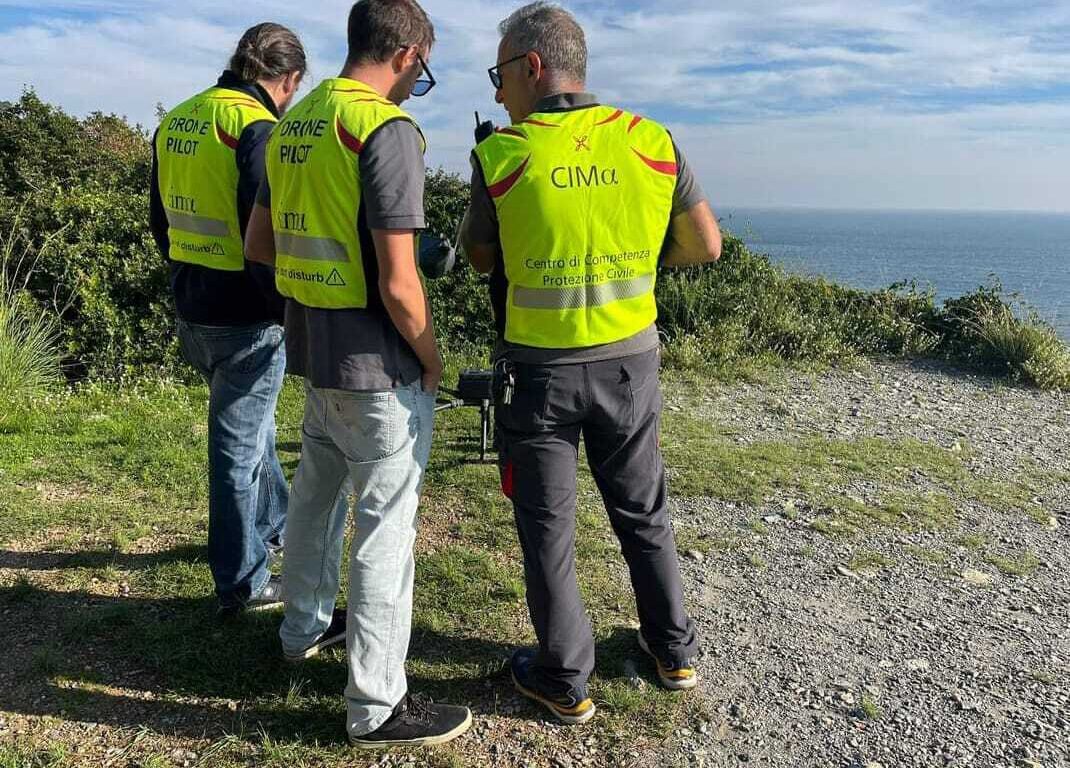 tre piloti di droni che si stanno coordinando durante l'esercitazione in zona collinare, in lontananza si vede il mare