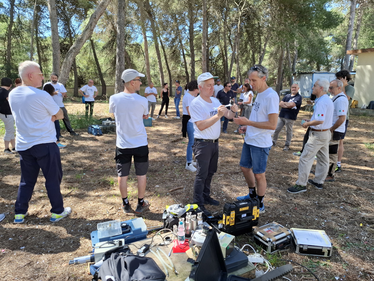 ricercatori in ambiente naturale, bosco, che parlano. Strumenti in primo piano. Altre persone sullo sfondo, e piante.