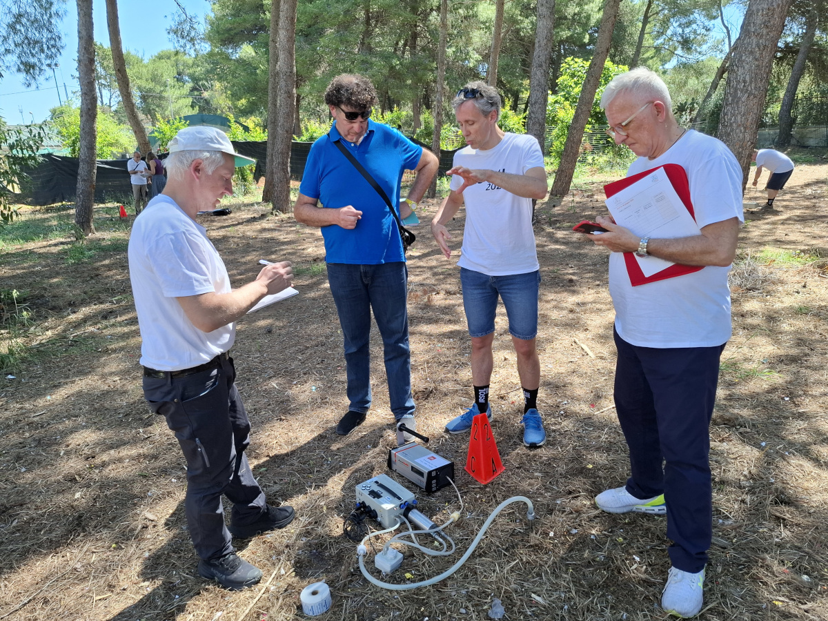 4 ricercatori in un bosco che parlano, strumentazione in primo piano ai loro piedi. Piante sullo sfondo.