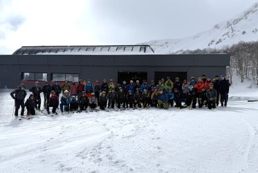 uomini e donne, vestiti da sci, davanti ad una stazione di un impianto di risalita in posizione per fare una foto. Sullo sfondo la stazione dell'impianto di risalita, le montagne innevate e il cielo terso. In primo piano la neve della pista da sci