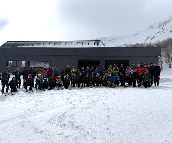 uomini e donne, vestiti da sci, davanti ad una stazione di un impianto di risalita in posizione per fare una foto. Sullo sfondo la stazione dell'impianto di risalita, le montagne innevate e il cielo terso. In primo piano la neve della pista da sci
