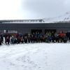 uomini e donne, vestiti da sci, davanti ad una stazione di un impianto di risalita in posizione per fare una foto. Sullo sfondo la stazione dell'impianto di risalita, le montagne innevate e il cielo terso. In primo piano la neve della pista da sci