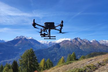 drone che vola, in secondo piano montagne, prato e alberi in primo piano, cielo azzurro.