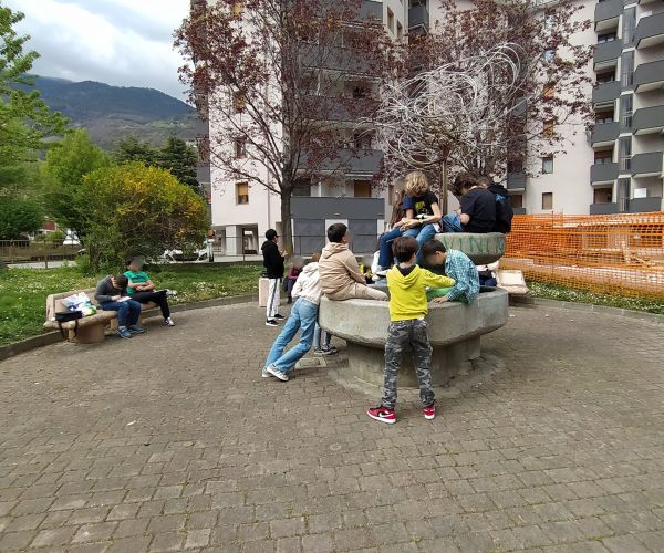 bambini e bambine sedute su un muretto che scrivono su un taccuino seguendo le indicazioni dell'esperto e delle professoresse, condomini sullo sfondo