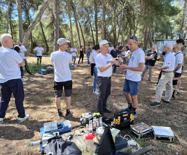 ricercatori in ambiente naturale, bosco, che parlano. Strumenti in primo piano. Altre persone sullo sfondo, e piante.