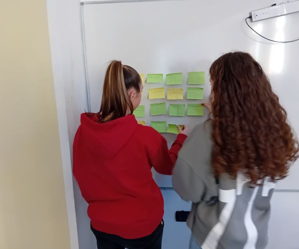 Due ragazze delle scuole superiori, girane di schiena, sistemano dei post it colorati e scritti sulla lavagna bianca