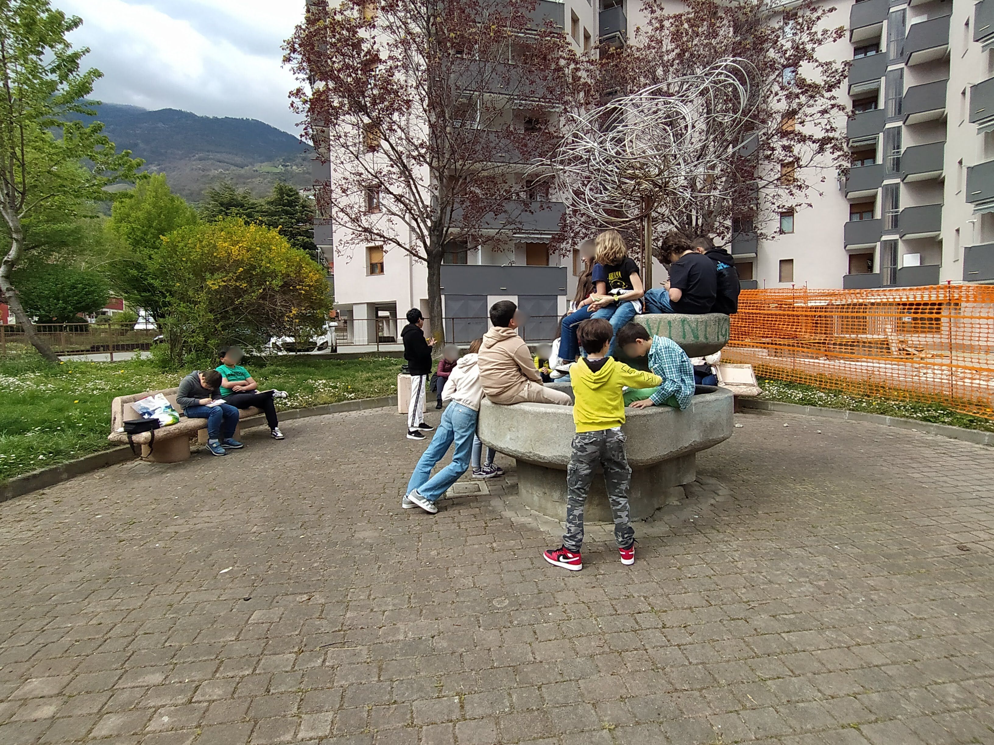 bambini e bambine sedute su un muretto che scrivono su un taccuino seguendo le indicazioni dell'esperto e delle professoresse, condomini sullo sfondo