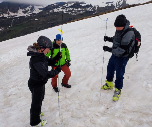 una donna e due uomini sulla neve, con sonde da valanga, che fanno misure. Sullo sfondo montagne