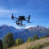 drone che vola, in secondo piano montagne, prato e alberi in primo piano, cielo azzurro.