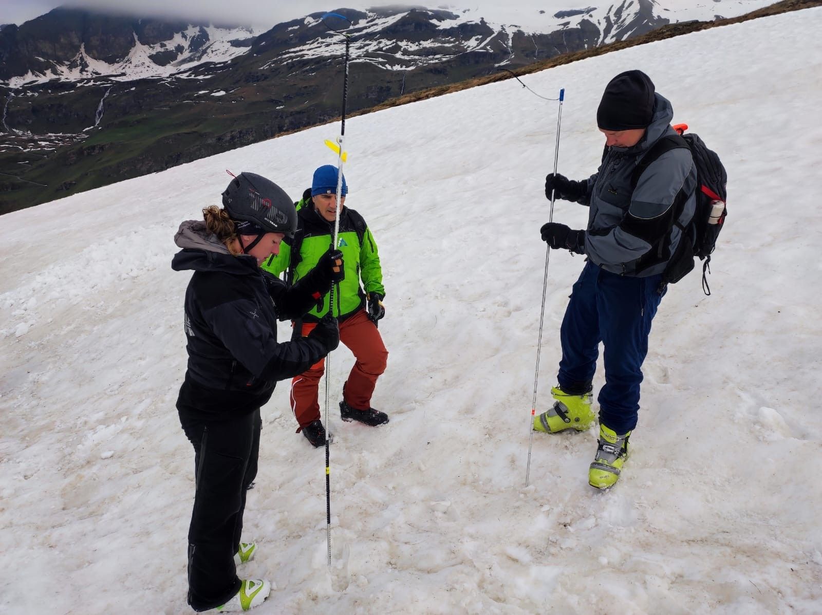 una donna e due uomini sulla neve, con sonde da valanga, che fanno misure. Sullo sfondo montagne